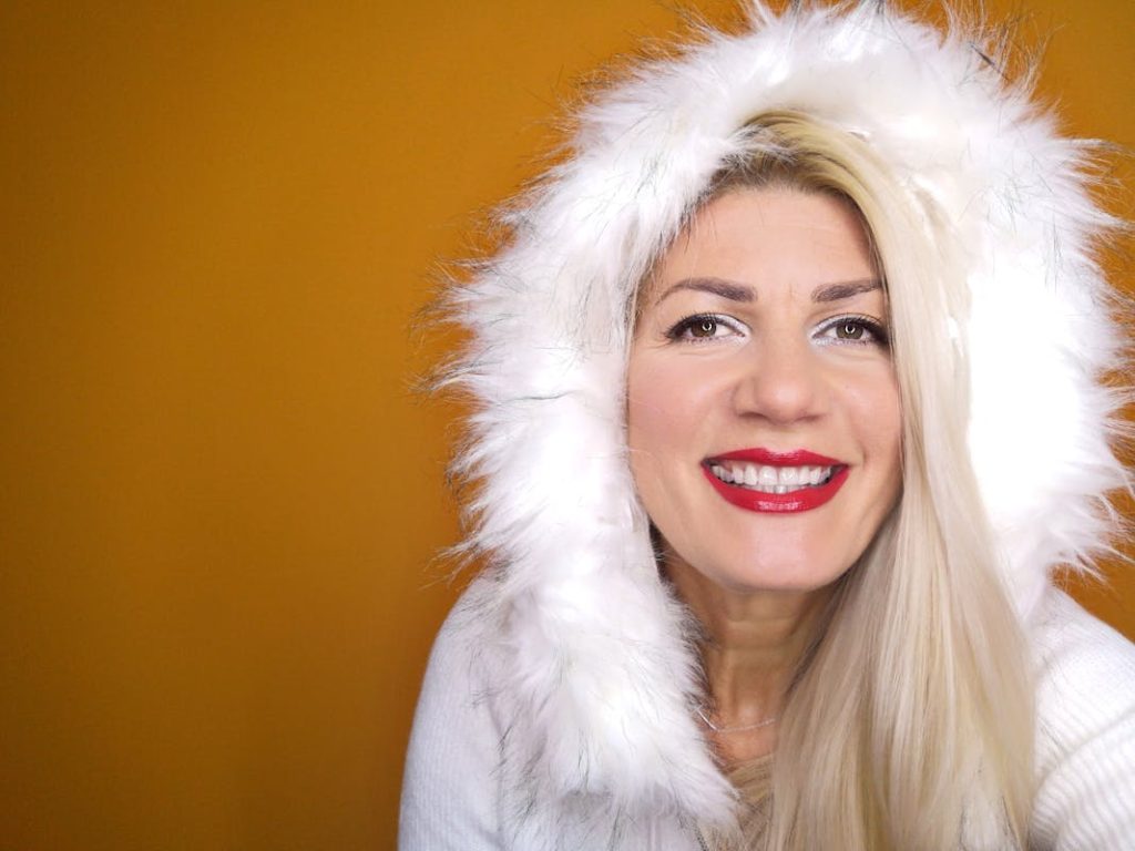 Cheerful woman wearing a white fur hood smiling against a vibrant orange backdrop.
